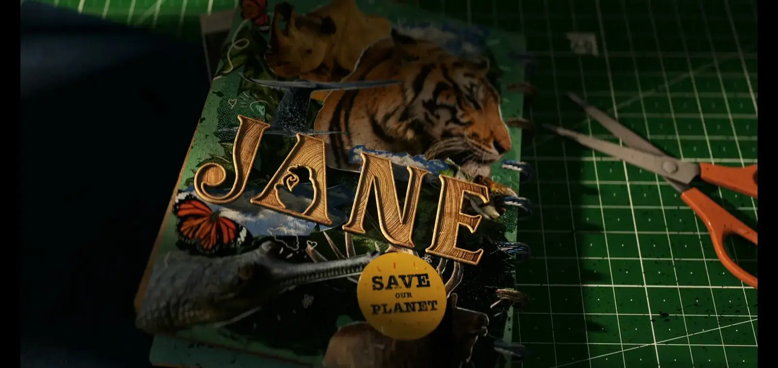 A colorful scrapbook or poster with tiger images, butterflies, and nature-themed decorations, with wooden letter cutouts spelling 'JANE' and a yellow circle sticker reading 'SAVE OUR PLANET,' on a green grid cutting mat with orange scissors nearby.