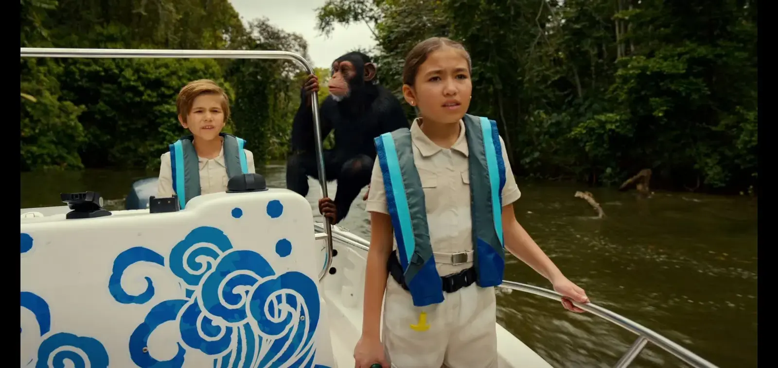 Three children and a chimpanzee on a boat, with a river and green trees in the background. The children are wearing life jackets and looking concerned, with the girl in front holding onto the boat railing.