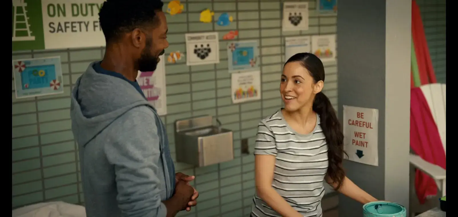A young woman with long dark hair, wearing a striped t-shirt, smiling and talking to a man with short black hair and a beard, wearing a gray hoodie, inside a colorful room with bulletin board, safety signs, and a bucket.