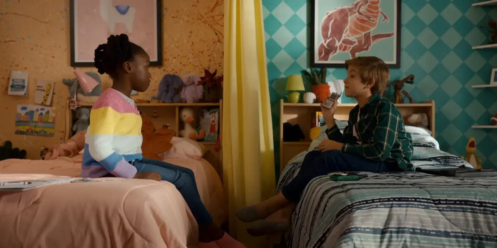 A girl and a boy sit across from each other on beds in a colorful bedroom, separated by a yellow curtain. The girl wears a pastel rainbow sweater, and the boy holds a remote control. The room has decorated walls with animal artwork, stuffed animals, books, and toys.