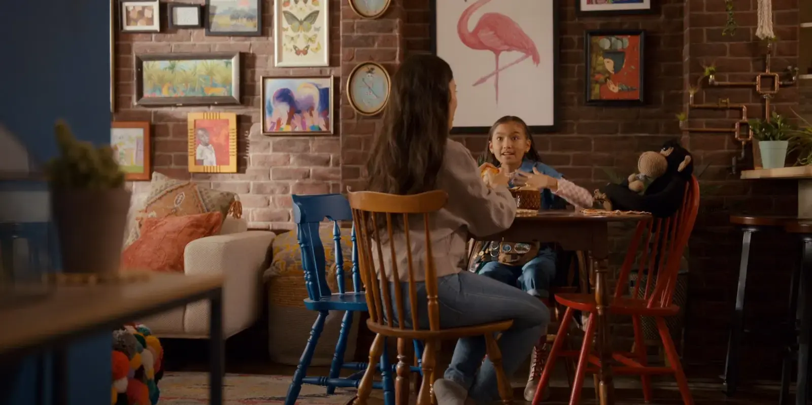 A woman and a girl are sitting at a round wooden table in a cozy cafe with a brick wall decorated with various framed artworks, including a flamingo, butterflies, and colorful paintings. The girl appears surprised or excited, while the woman is turned towards her, engaging in conversation. There are