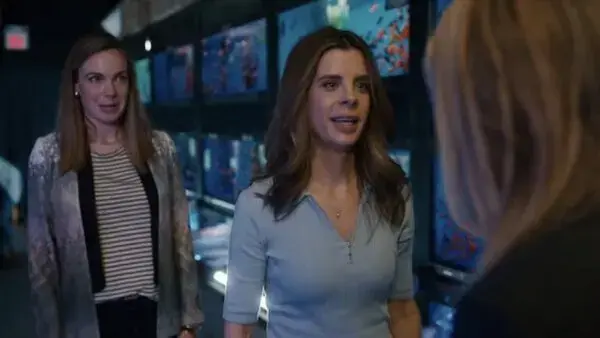 Two women are having a conversation in what appears to be an electronics store or aquarium shop, with aquariums or screens displaying colorful fish in the background.