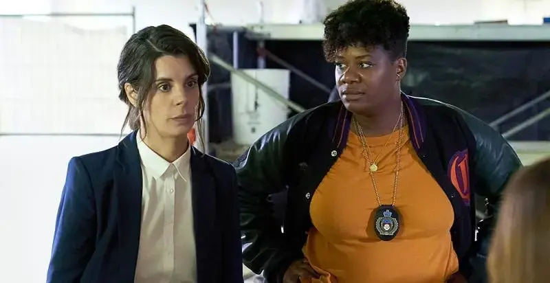 Two women standing together, one in a navy blazer and white shirt, the other in an orange shirt with a police badge necklace, in an industrial or warehouse setting.