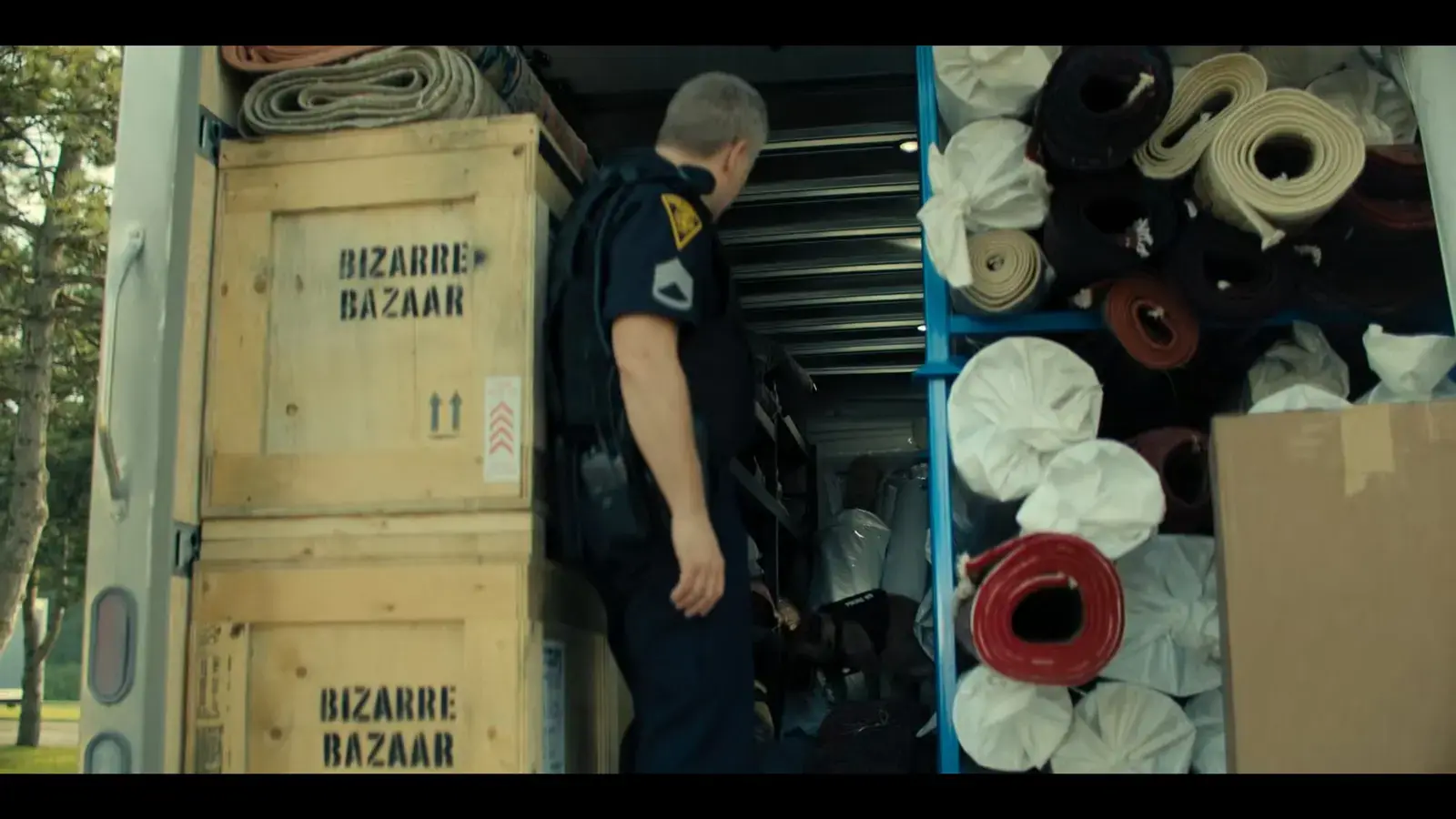 A person in a police uniform with a backpack looking inside a truck filled with rolled-up mats and boxes labeled 'Bizarre Bazaar'.