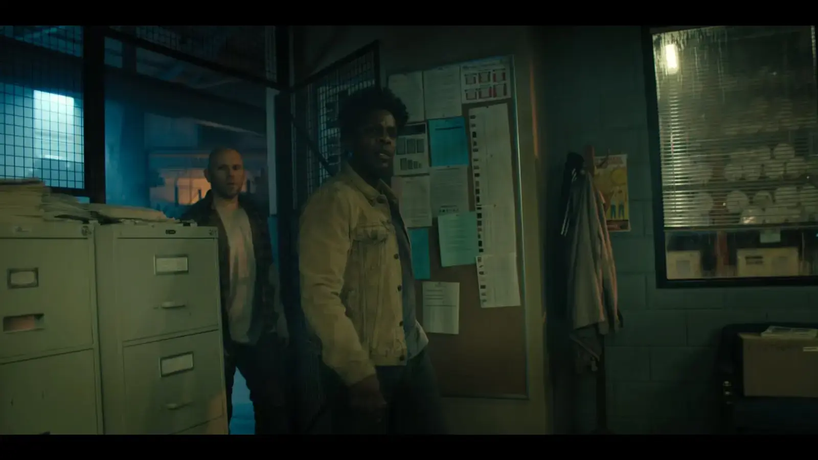Two men standing in a dimly lit office with file cabinets, bulletin board, and window with blinds.