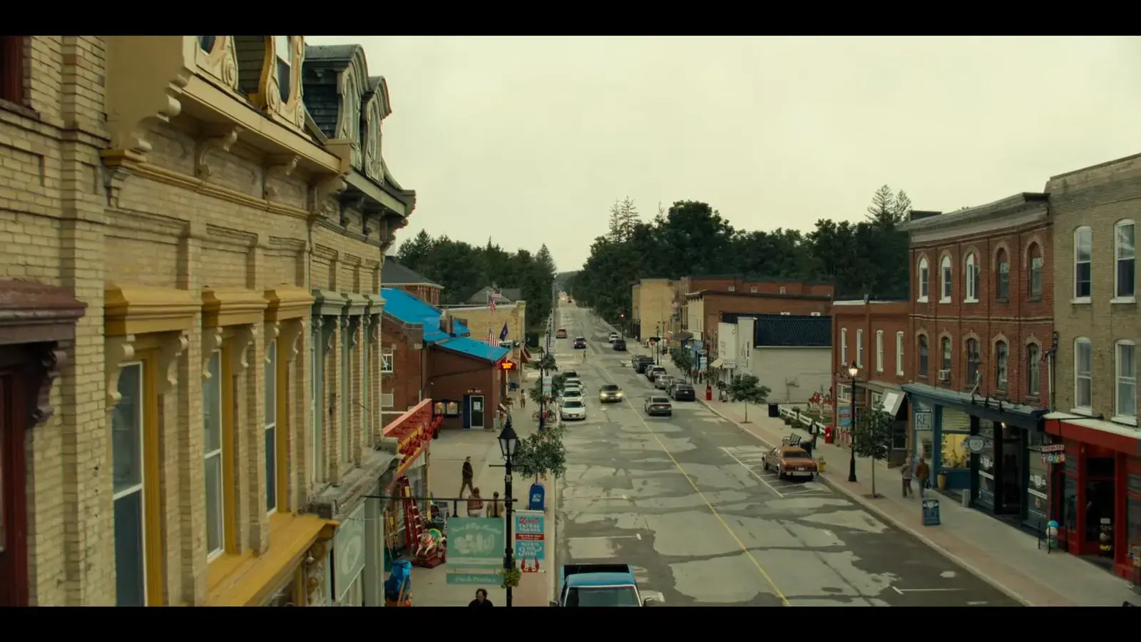 View of a small town main street with brick and stone buildings, some with colorful facades, storefronts, flower baskets, trees, parked cars, and a few pedestrians, under a cloudy sky.