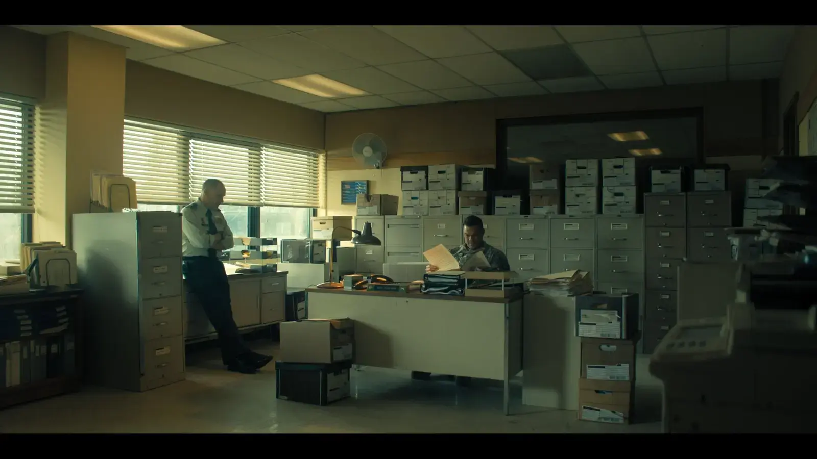 An office scene with two men, one standing and one sitting, surrounded by filing cabinets, boxes, and office supplies, with sunlight coming through window blinds.