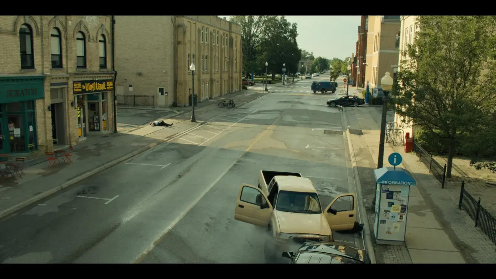 A beige SUV has crashed into a parking meter at an intersection, with its driver’s side door open. There is smoke coming from the front of the SUV. Nearby, a person lies motionless on the sidewalk, and other cars are visible on the street.