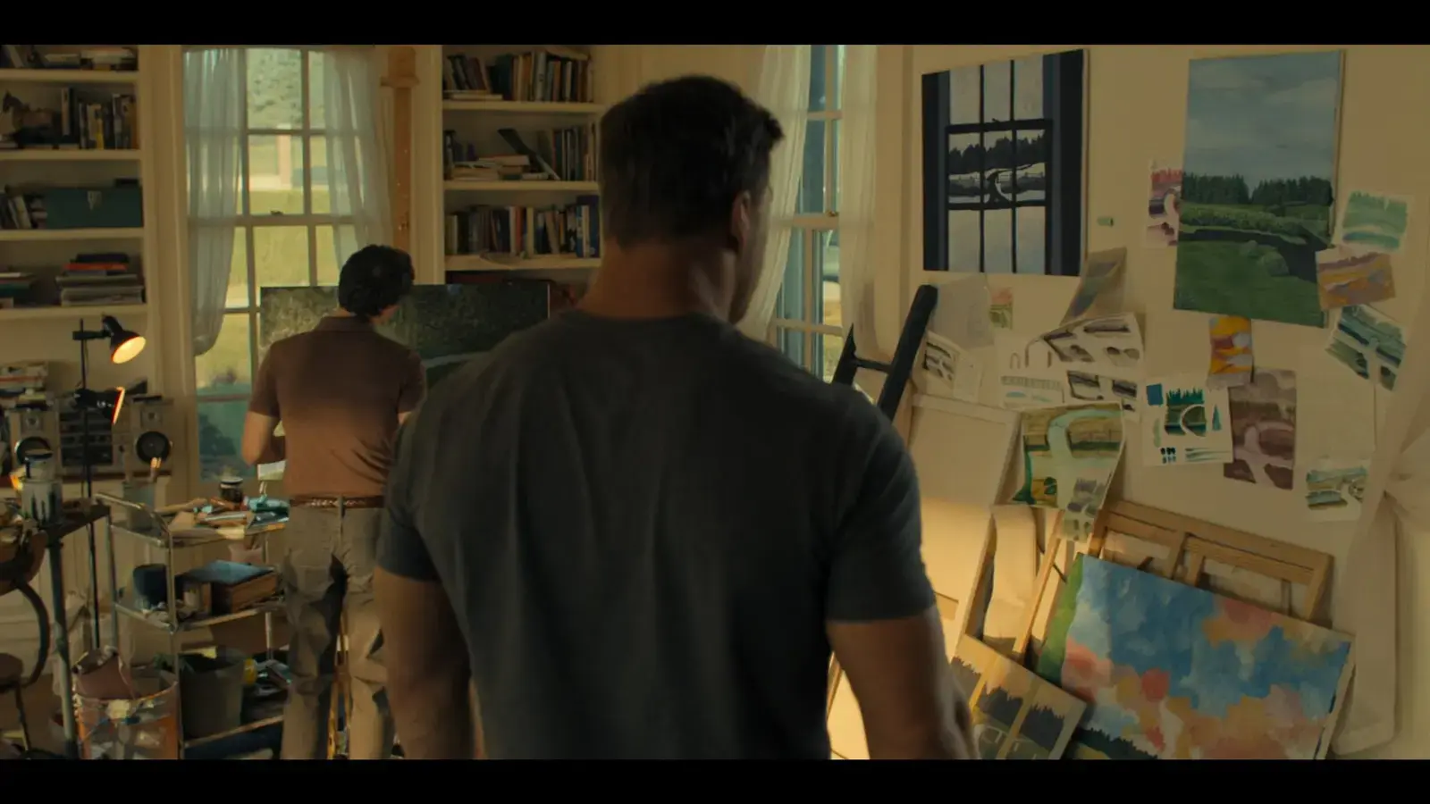 Two men working in an art studio with paintings, sketches, and art supplies. One man is in the foreground looking at the art while another is in the background working on a painting.