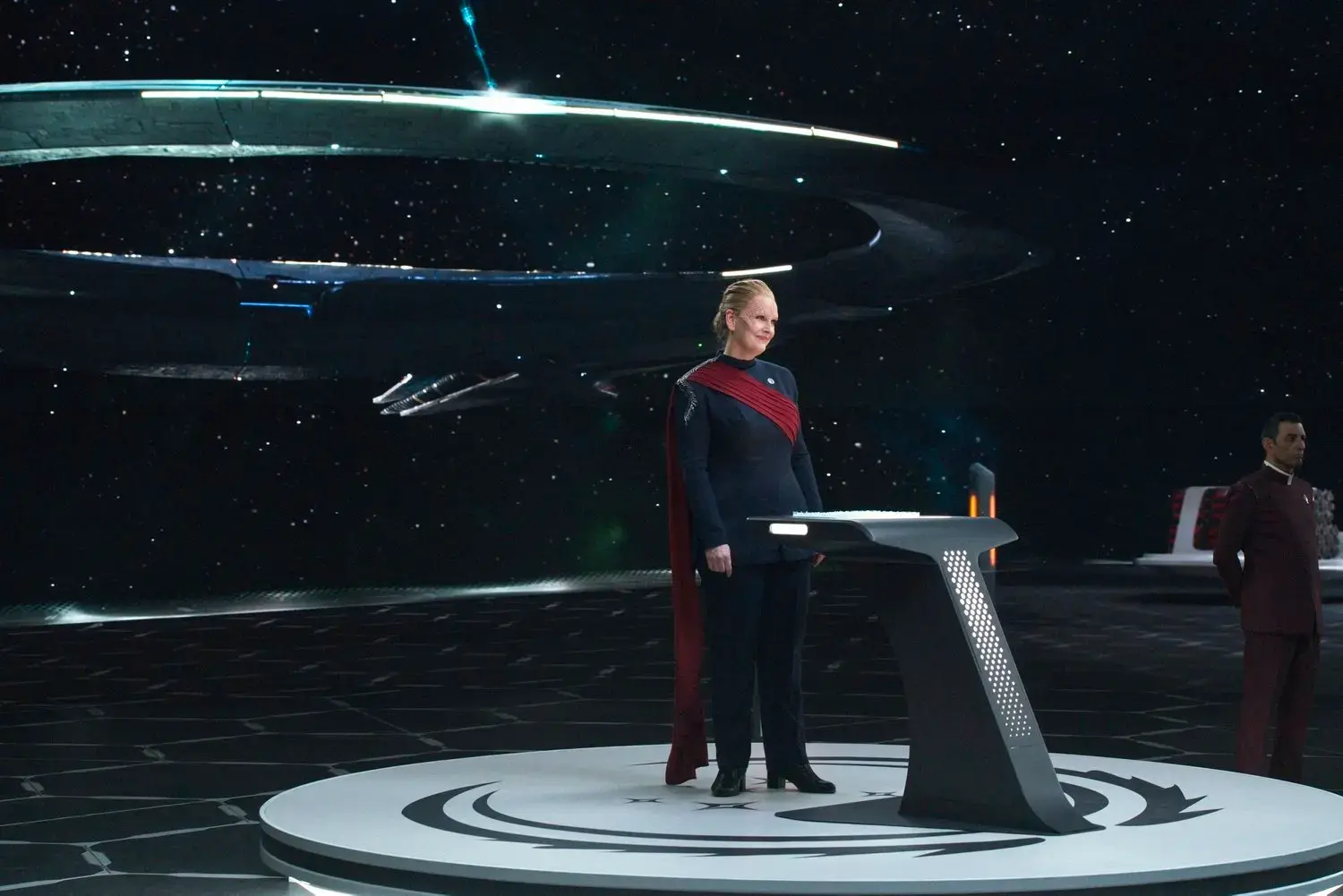 A woman in a dark uniform with red accents stands at a futuristic podium on a spaceship or space station set against a backdrop of stars and a large spaceship in space.