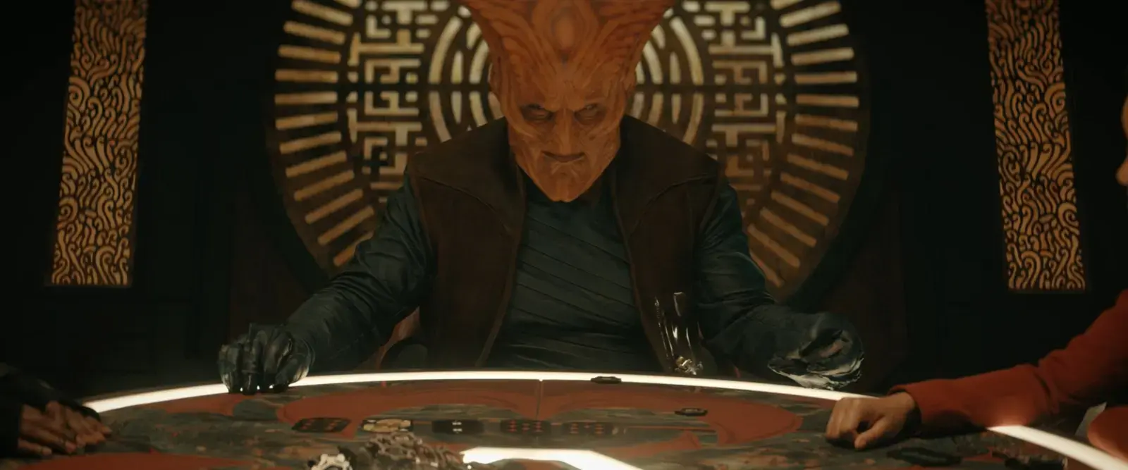 A humanoid character with an orange, textured alien face and large forehead, seated at a gaming table with poker chips, against a decorative background with Asian-inspired patterns.