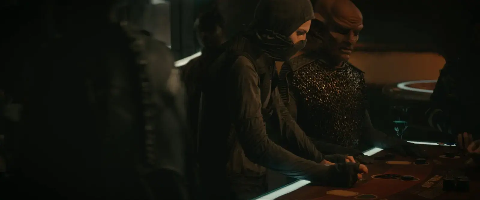 People in dark clothing playing poker at a casino table in a dimly lit room.