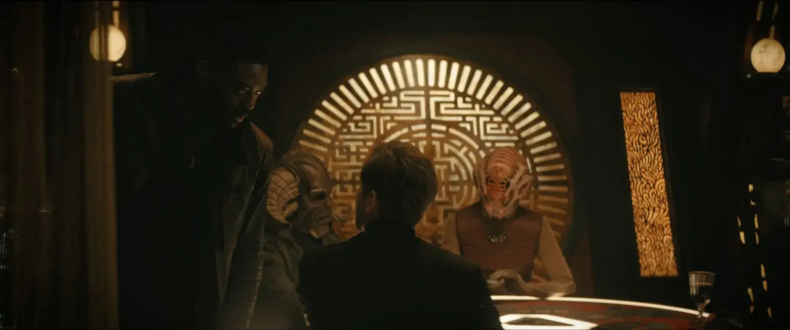 A group of people, some with alien or creature-like appearances, are gathered around a table in a dimly lit room with Asian-inspired decor and intricate backlit designs.