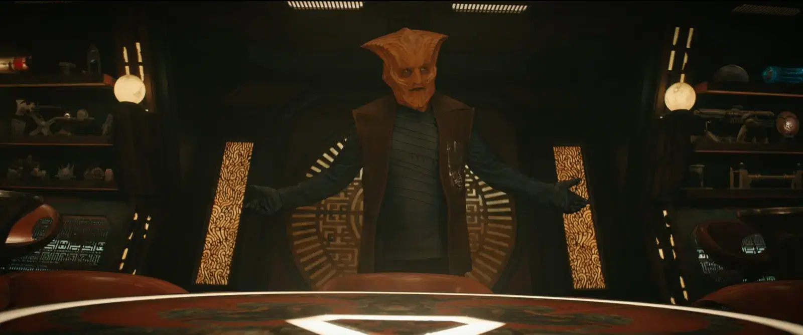 A character with an alien-like face and orange skin stands with arms outstretched in a dark room with shelves of objects and glowing panels.