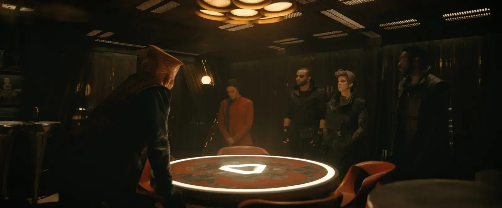 Scene in a dimly lit room with a circular table at the center, featuring the Assassin's Creed logo. Five people stand around the table, one person with a hooded cloak and others in tactical clothing, suggesting a meeting or planning session.