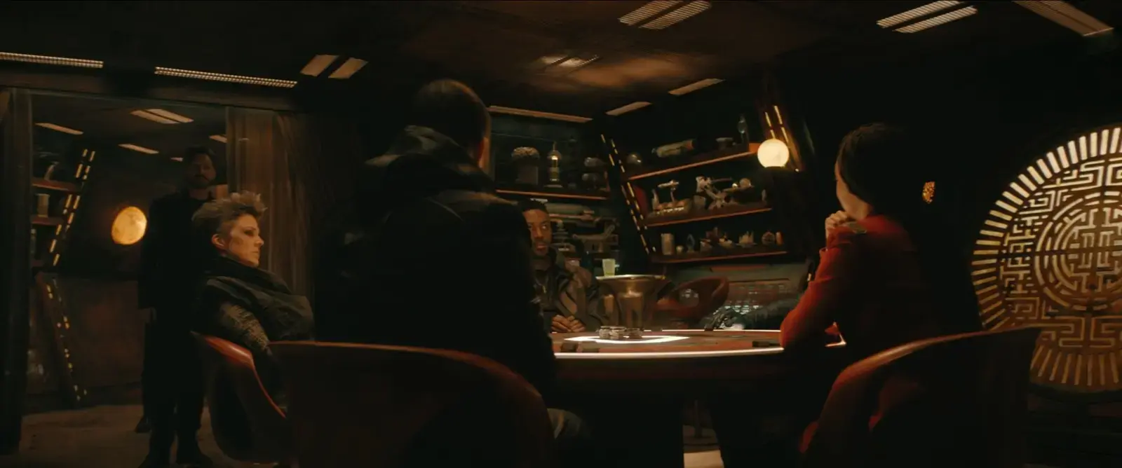 Scene from a sci-fi movie showing a group of people seated around a circular table in a dimly lit room with shelves containing various artifacts and symbols in the background.