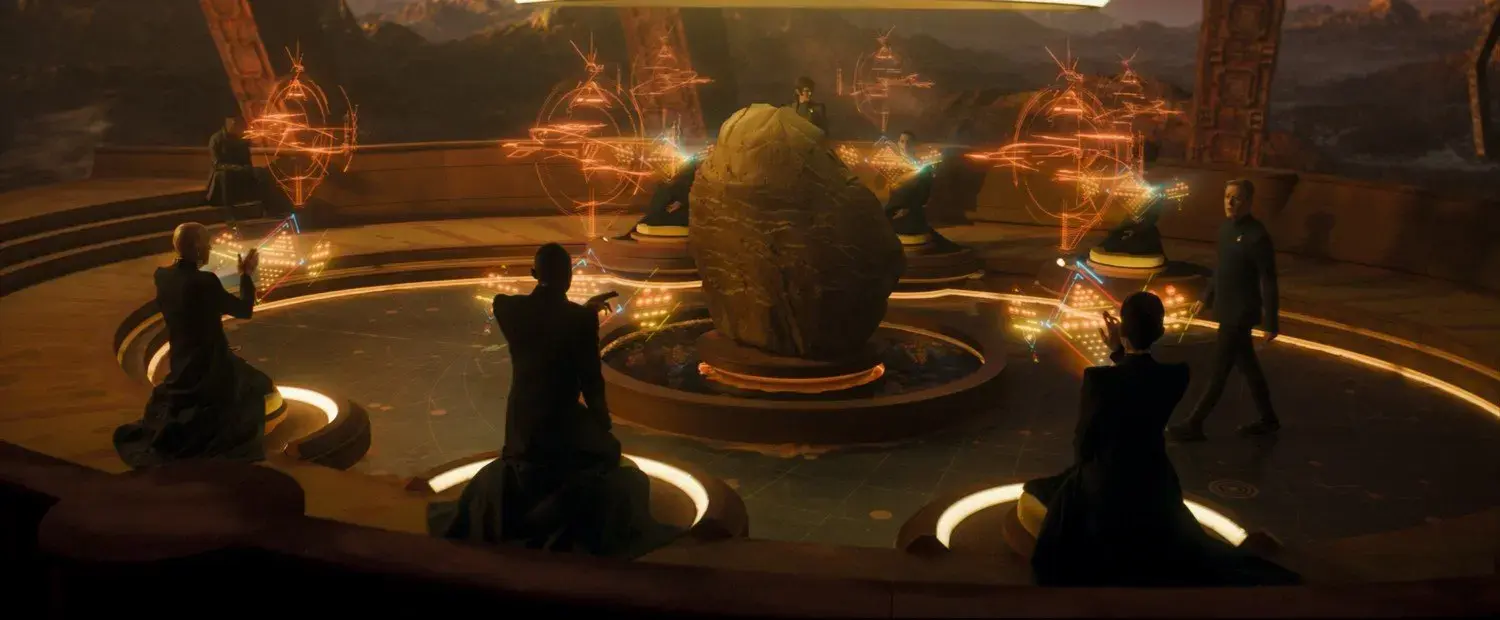 A scene from a futuristic meeting room with seven individuals, some sitting and some standing, around a round table with a large rock at the center. The room features holographic displays with glowing orange symbols and a backdrop of mountains.