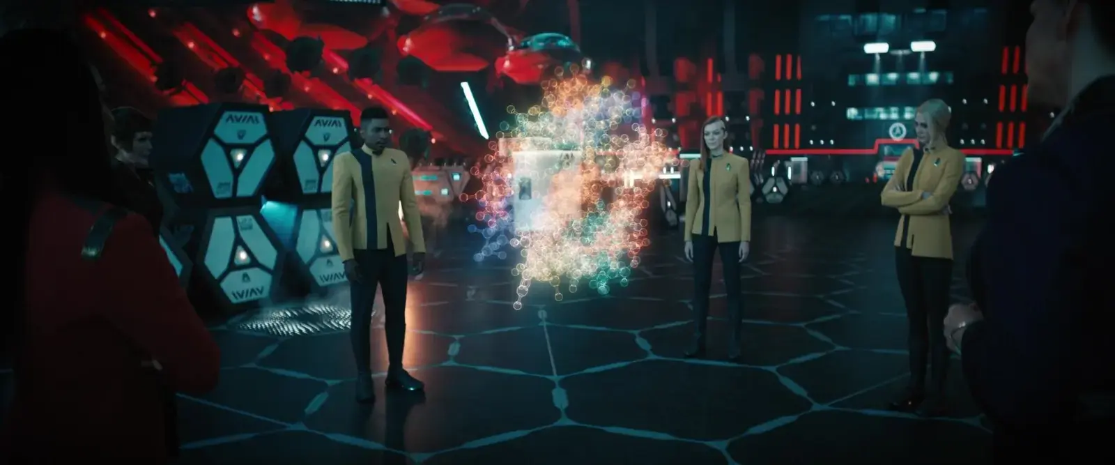 People in science fiction uniforms standing around a glowing holographic display in a high-tech spaceship or control room with futuristic equipment and red lighting.