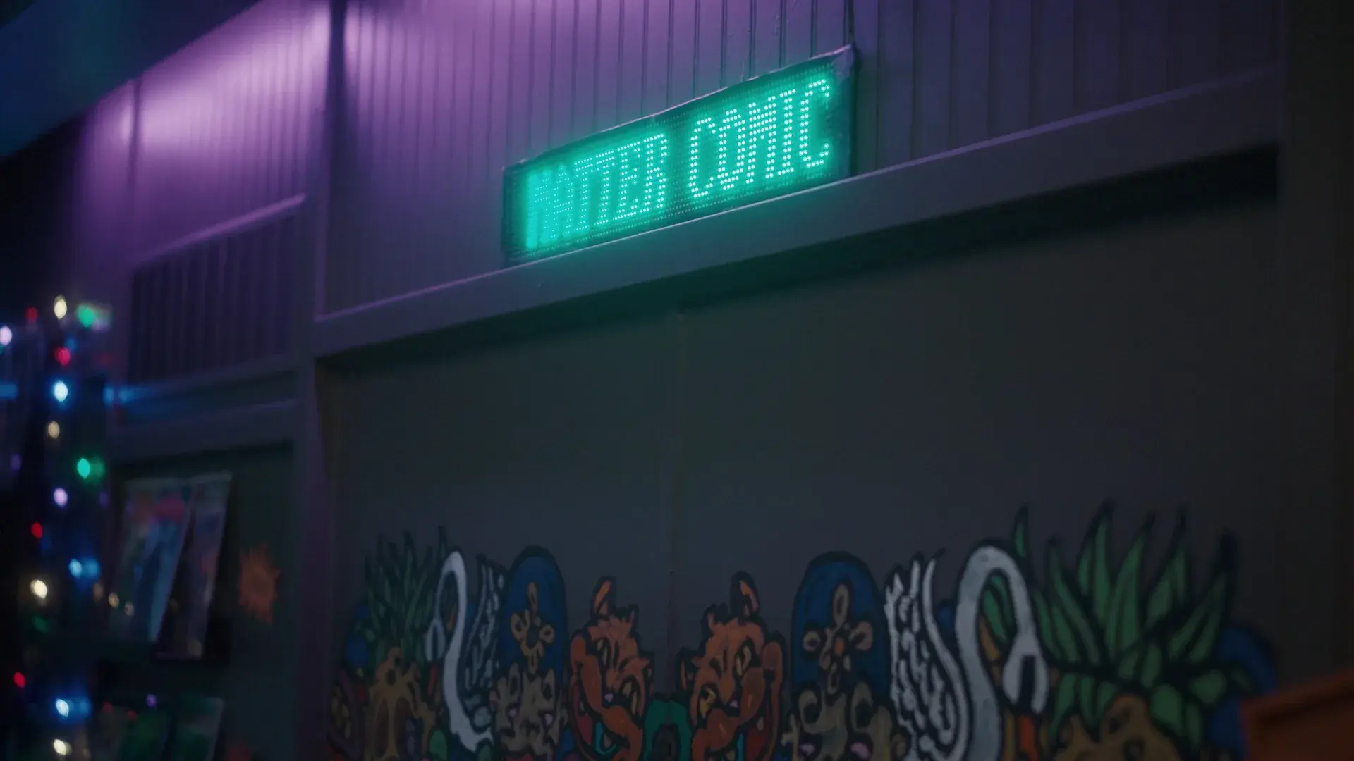 A comic shop at night under a glowing green neon sign, its wall covered in hand-painted monster murals