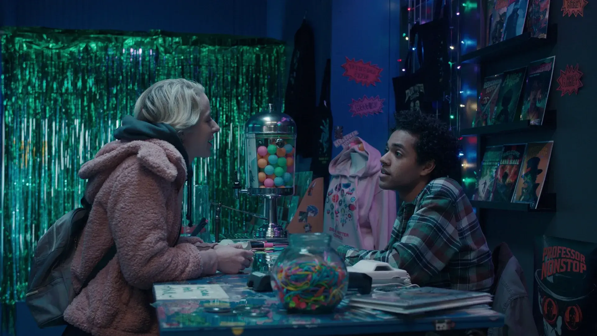 Two characters talk across the comic shop counter, surrounded by comics, candy jars and fairy lights