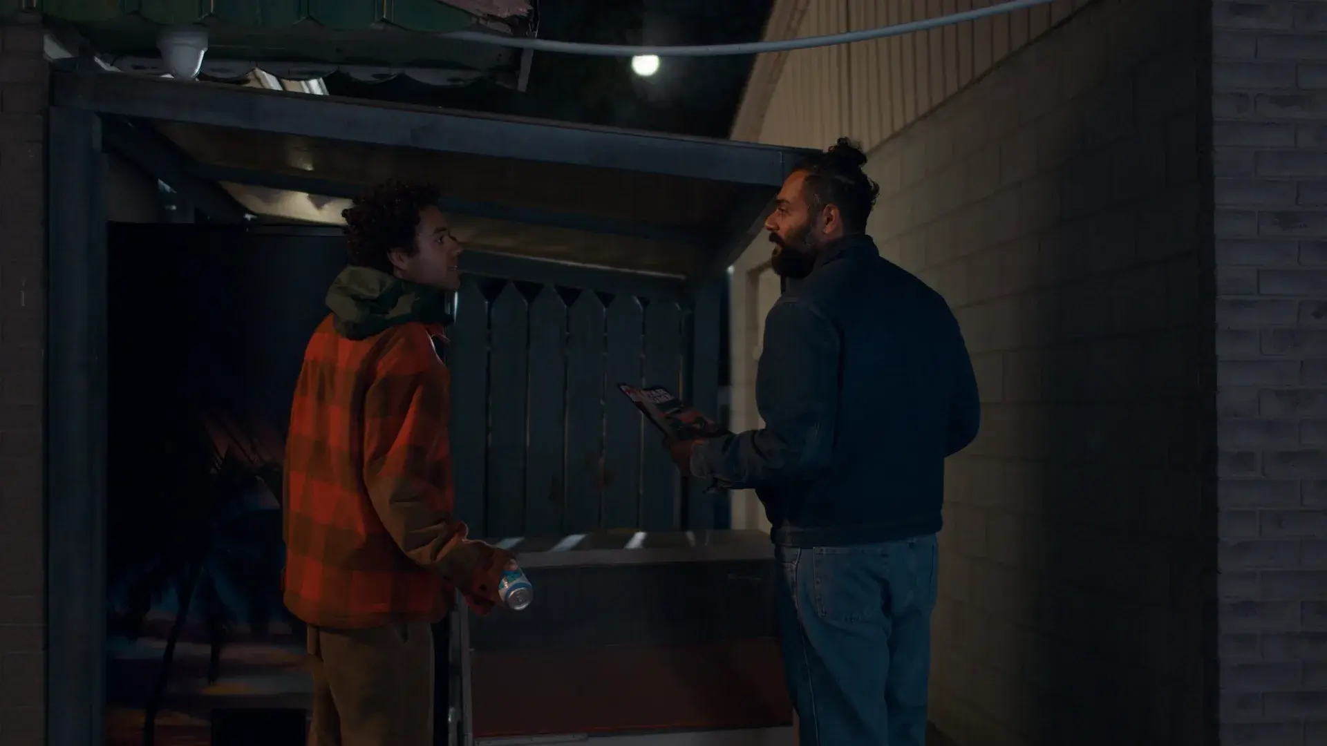Two men talk at night outside a wooden shed
