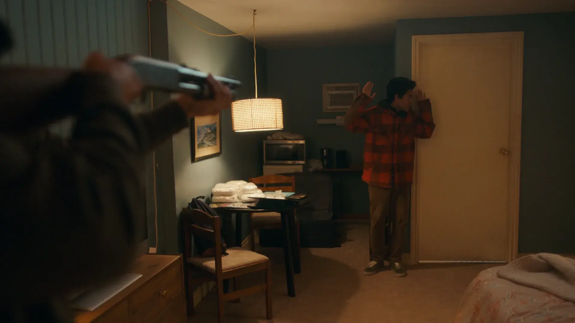 A man with a shotgun confronts another with his hands raised in a motel room
