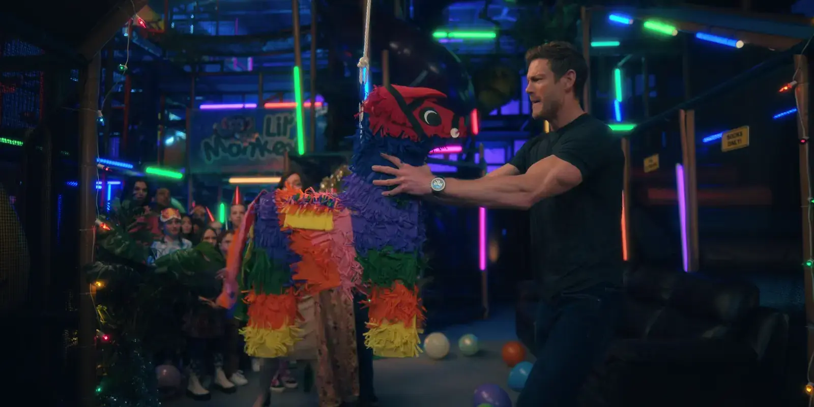 A man in a black t-shirt holding a colorful piñata shaped like a llama at an indoor party with neon lights and balloons, and group of children watching.