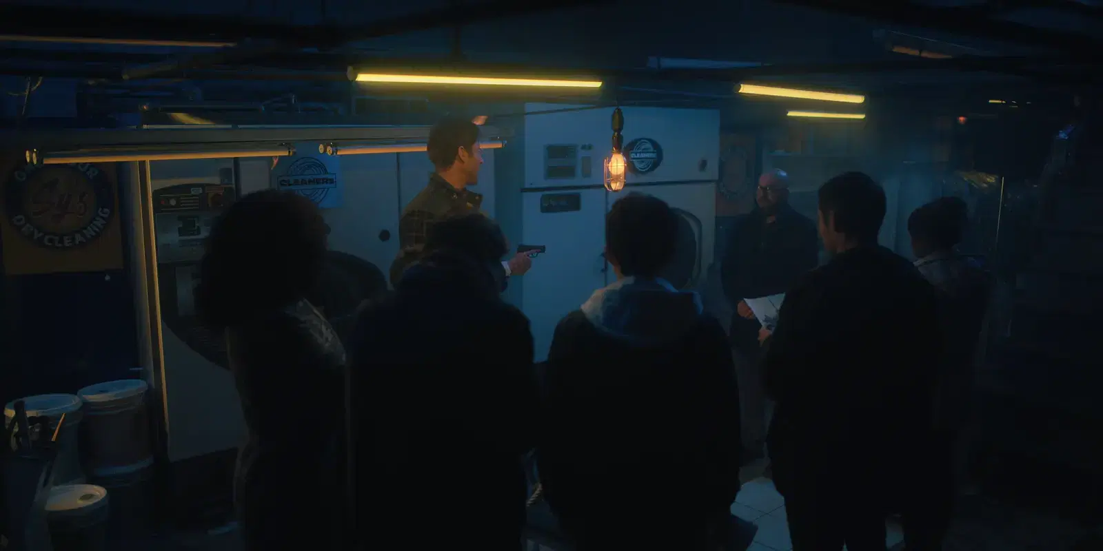 A small group of people in a dark laundry room, with two washers or dryers in the background, listening to a man speaking and holding a gun, with one person holding a paper.
