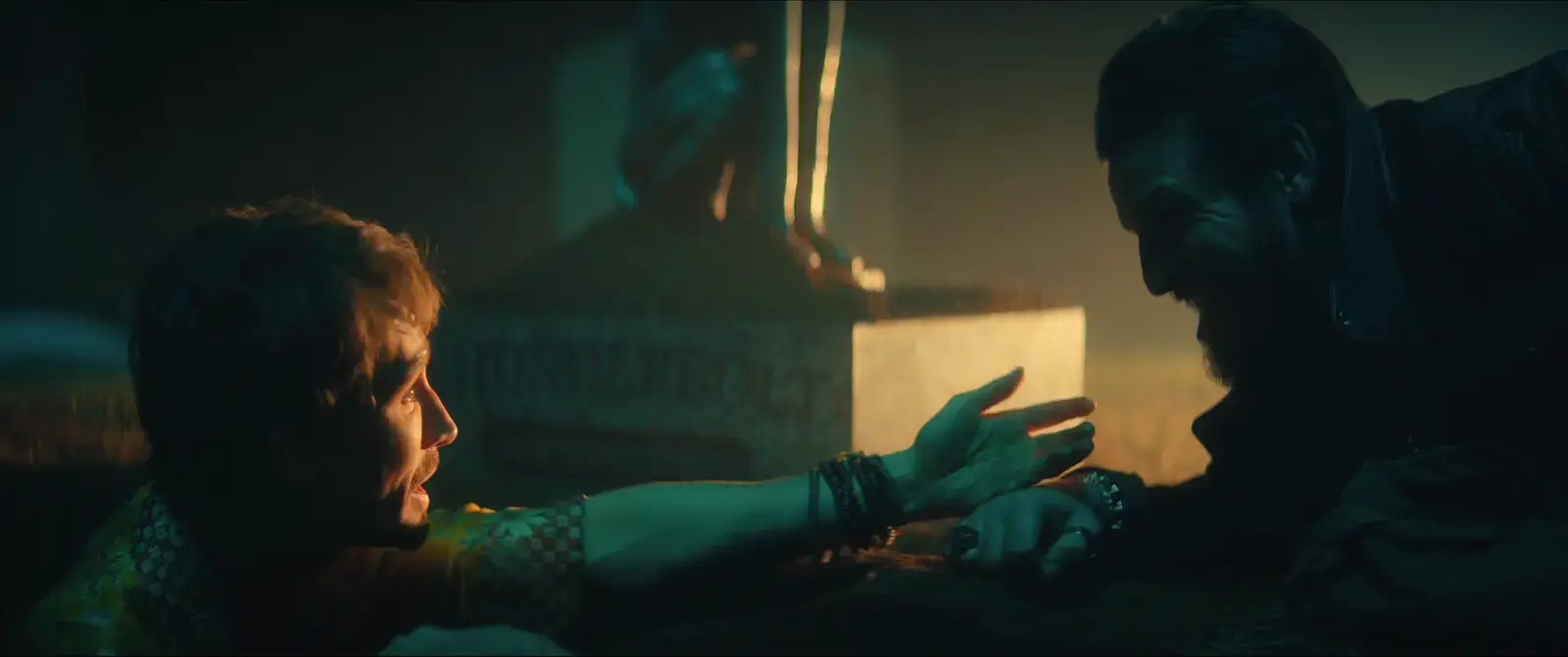 Two men are on the ground, one reaching out to the other in a dark, intense scene with dramatic lighting, with a statue or sculpture visible in the background.