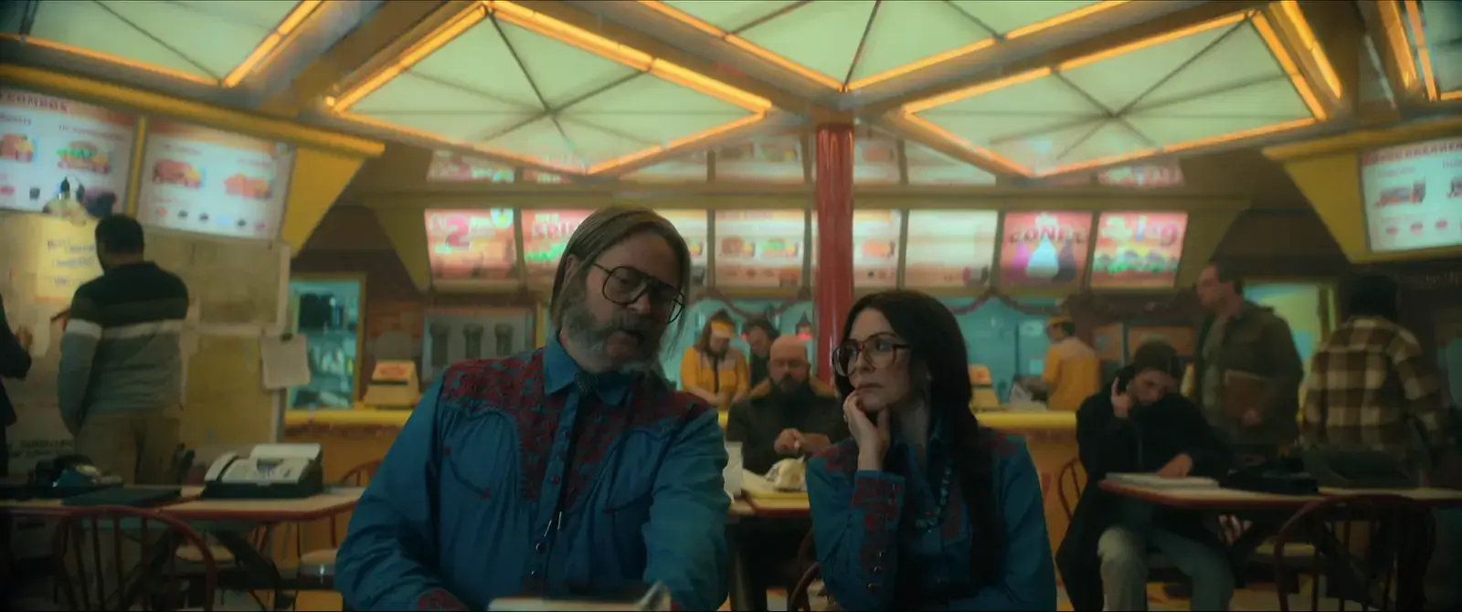 People sitting in a brightly lit fast-food restaurant, two main characters in focus talking, colorful menu signs overhead, others in the background.