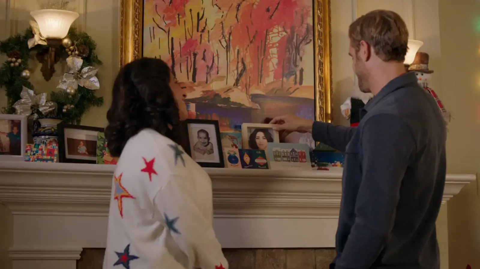 A man and woman standing near a fireplace decorated with Christmas wreaths, ornaments, and framed photos, with a large colorful painting of a forest scene hanging above.