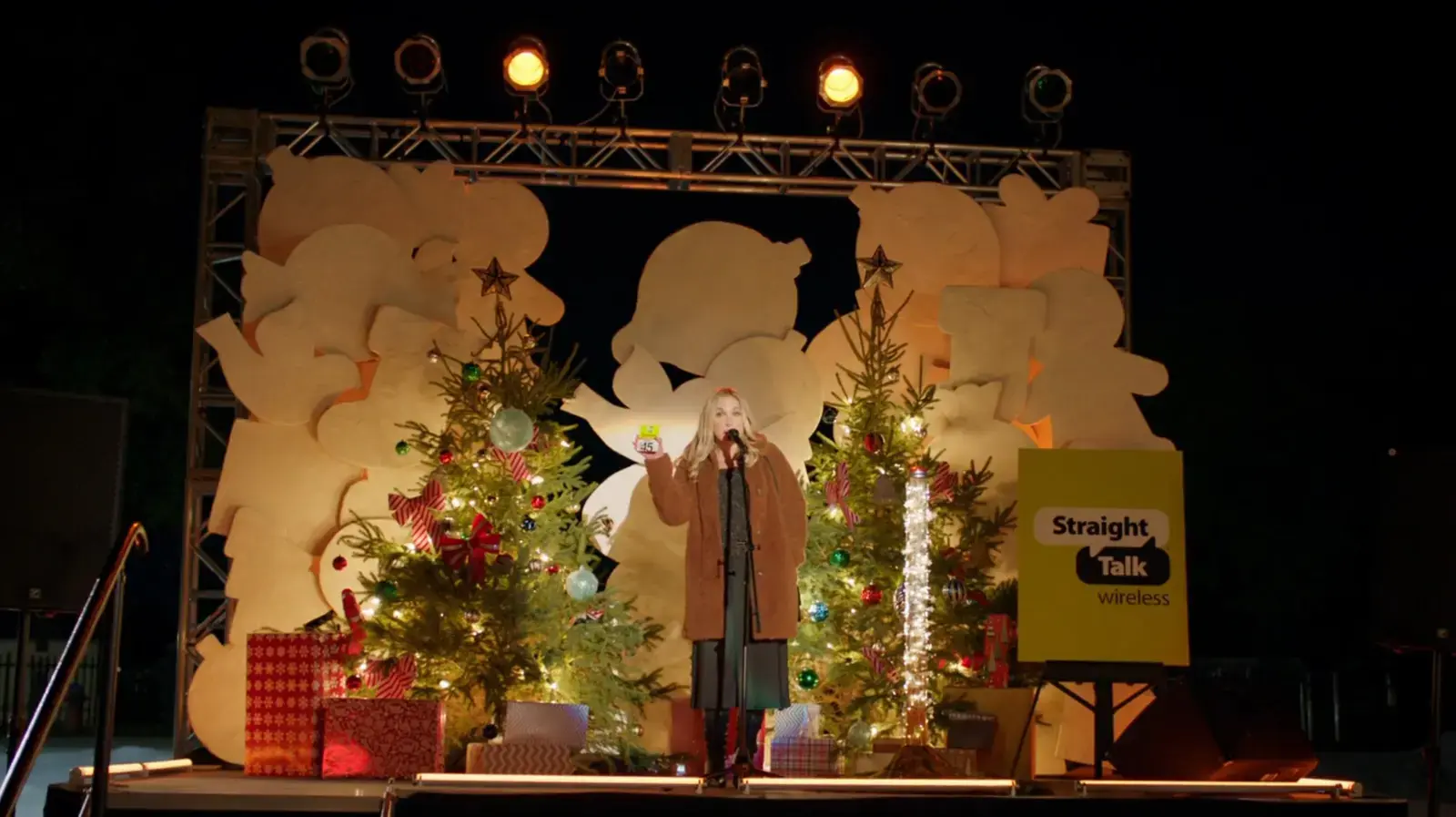 A woman speaking on stage at night with Christmas trees and decorations, large cut-out snowflakes in the background, and holiday presents nearby. There are stage lights overhead and a sign that reads 'Straight Talk wireless'.
