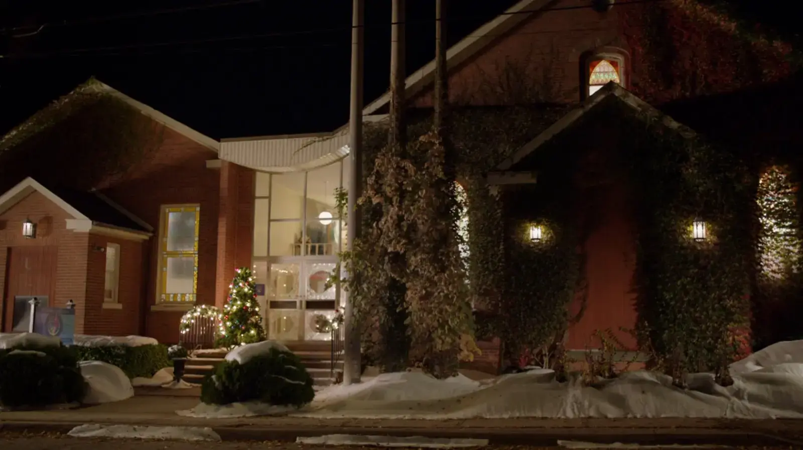 A house decorated for Christmas at night, with a Christmas tree illuminated outside, snow on the ground, and warm lights inside visible through large windows.