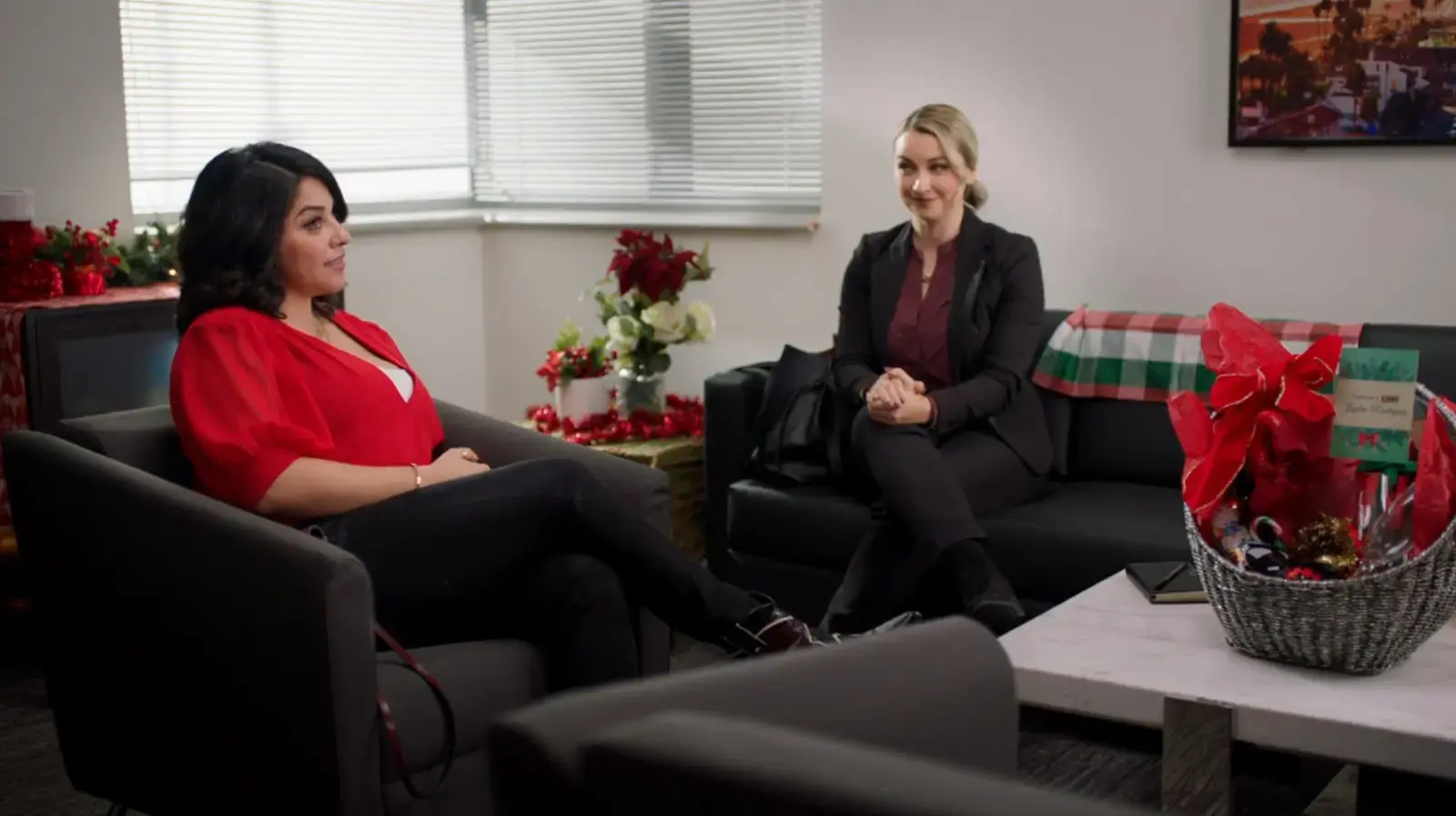Two women sitting on black sofas in an office decorated for Christmas, with poinsettias, a gift basket, and holiday decorations on the table and in the background.