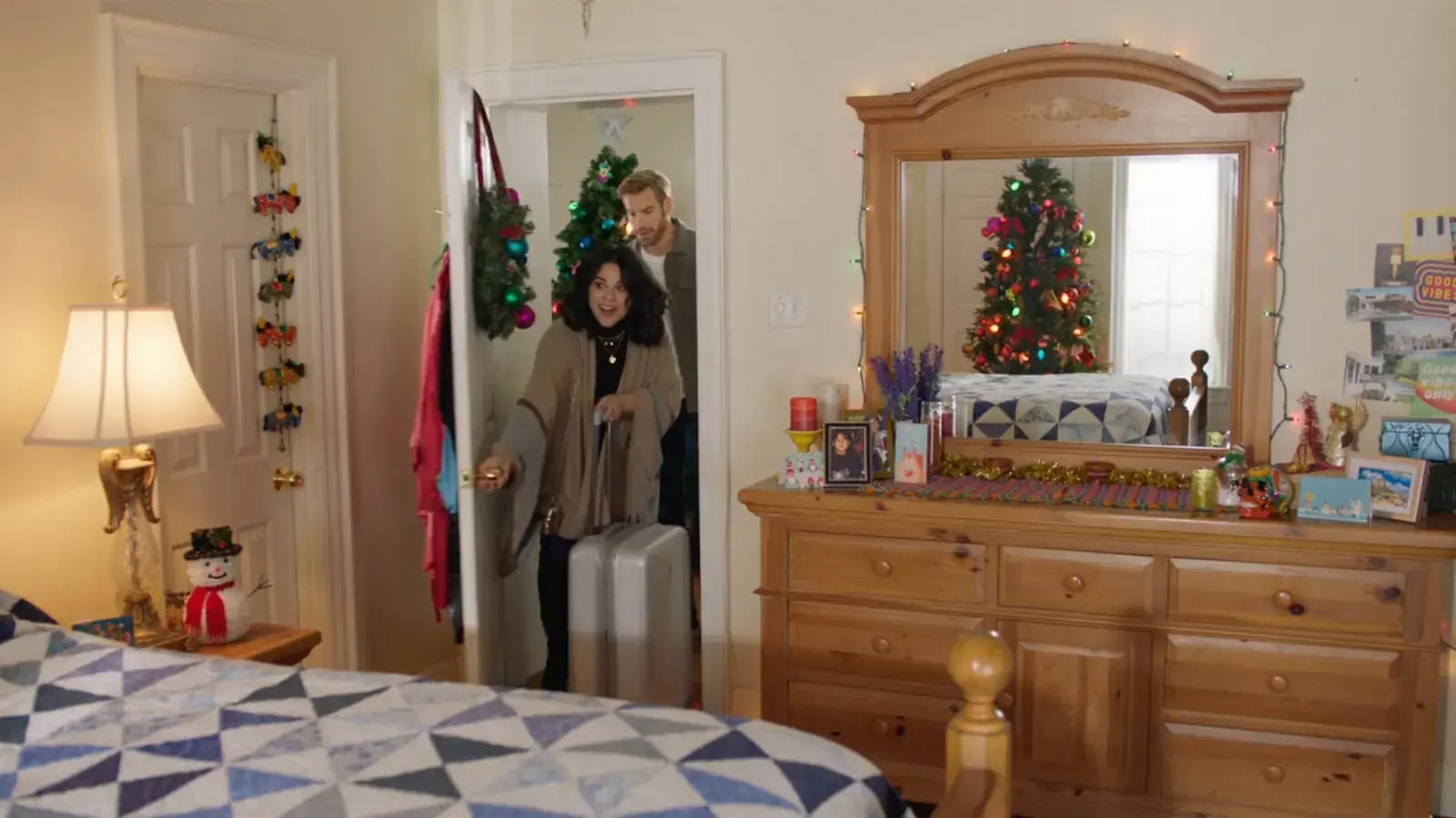 A woman and a man entering a bedroom decorated for Christmas. The woman is carrying a suitcase, and they are both smiling. There is a decorated Christmas tree in the reflection of a large wooden mirror on the dresser. The room has festive decorations including string lights, a snowman figurine, and 